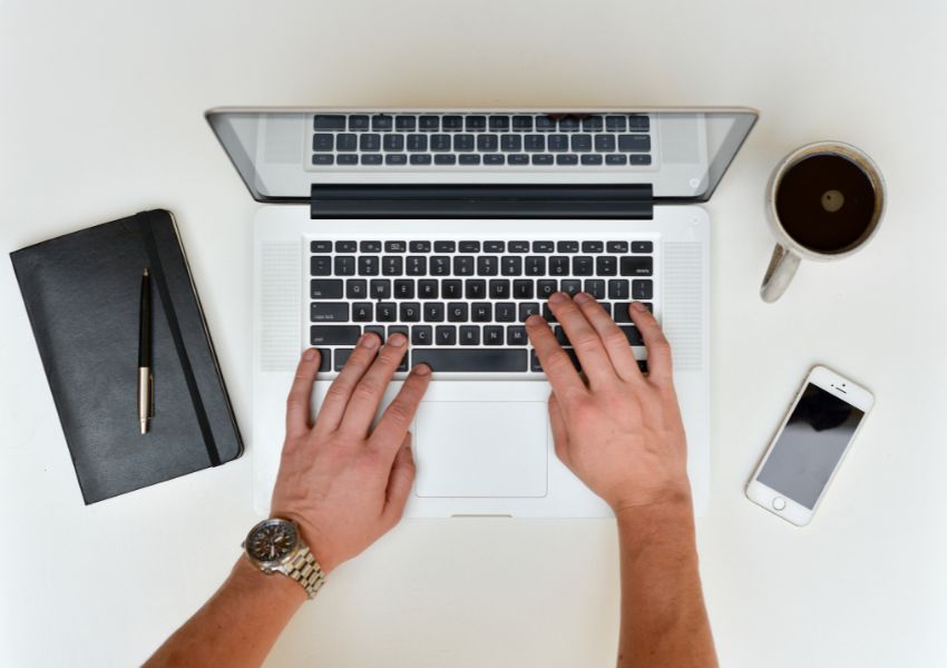 two hands typing on a laptop with a cup of coffee and notebook next to them